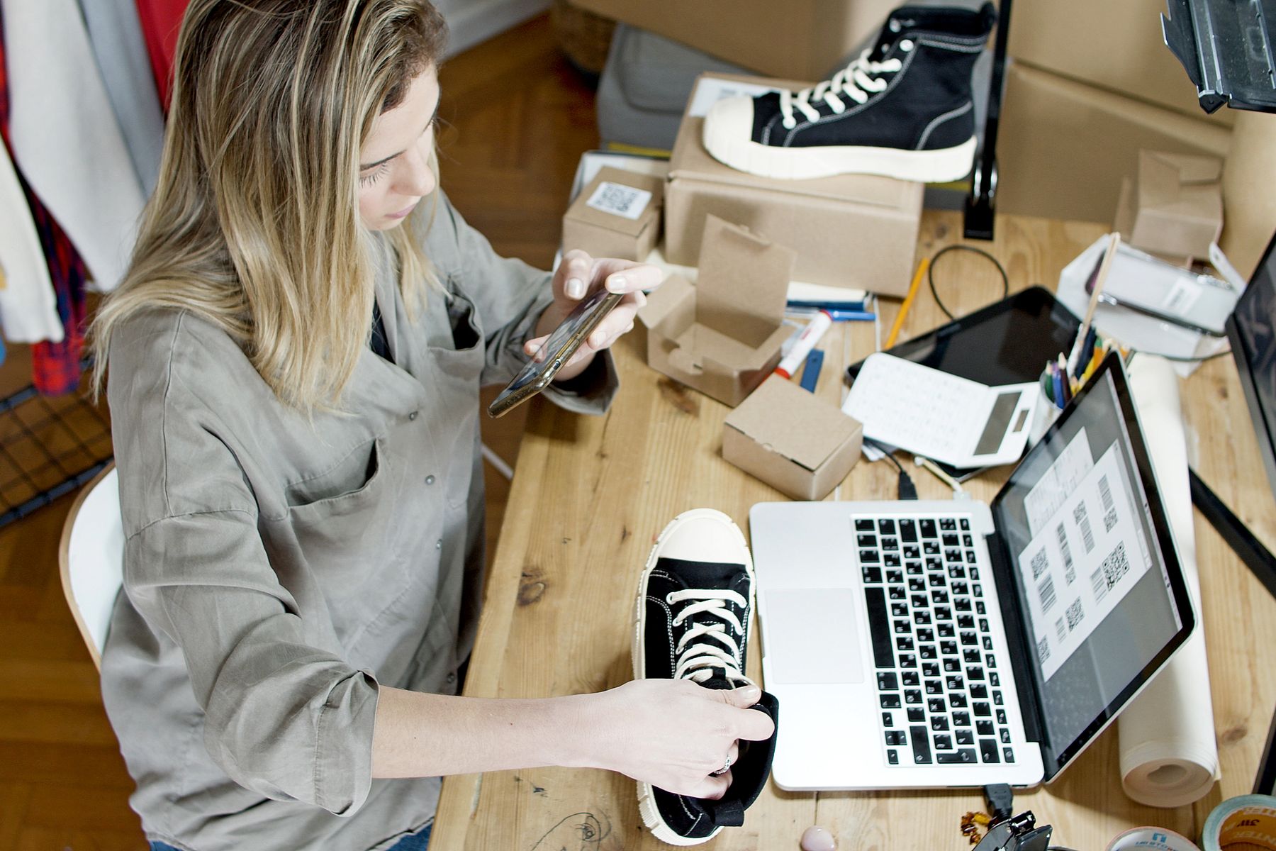 Emprendedora fotografía unas zapatillas con el móvil para venderlas online, junto a su portátil y cajas de envío